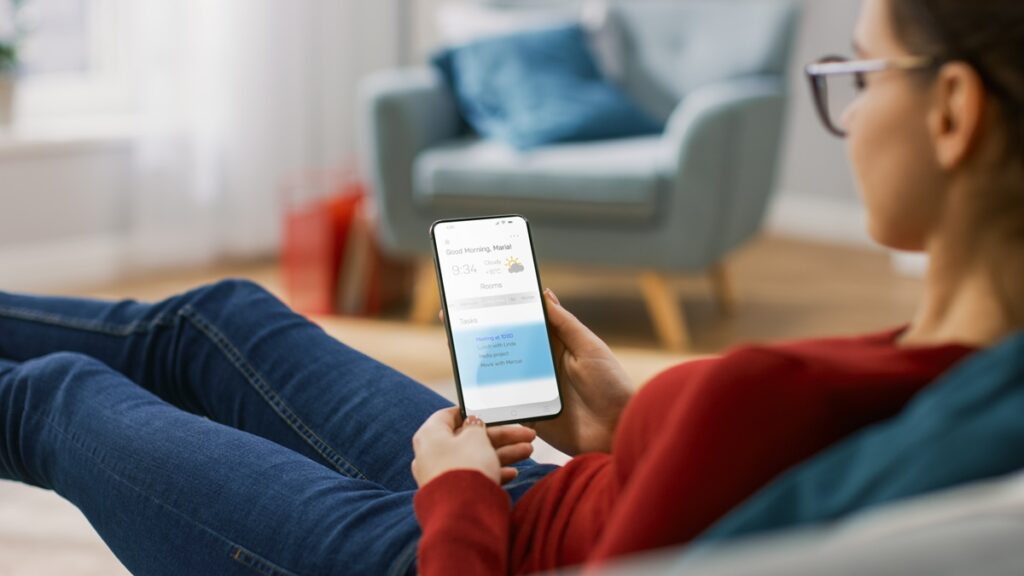 Mulher segurando celular com app de previsão do tempo, sentada no sofá