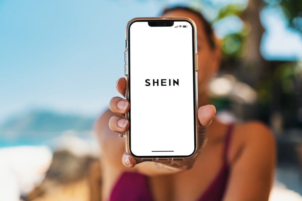 Mulher segurando celular na praia com logo Shein; pontos Shein